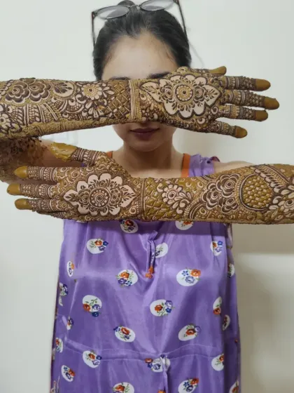 A happy client showing off her full bridal mehandi. The design is symmetrical on both hands, featuring a central mandala on the back of the hand and detailed patterns extending up the arms.