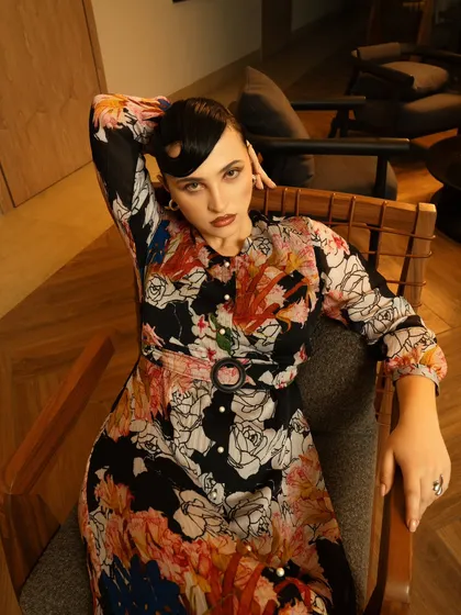 A relaxed, seated portrait that highlights a floral print dress. The warm indoor lighting and comfortable setting make the fashion feel accessible and stylish.