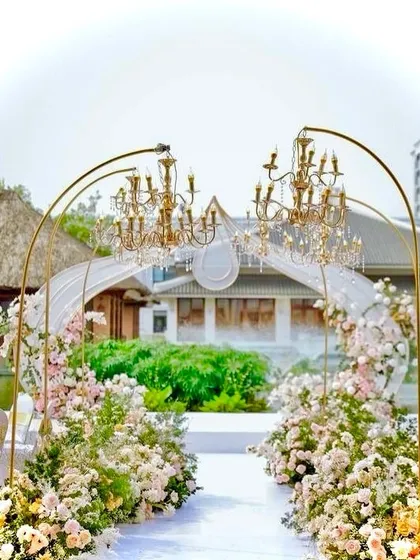 A beautiful and romantic wedding aisle created with golden arches holding elegant chandeliers. The path is lined with lush arrangements of pastel flowers.