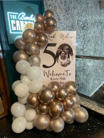 A personalized welcome sign for a 50th birthday party. The sign features a photo of the couple and is framed by a chic white and rose gold balloon arrangement.