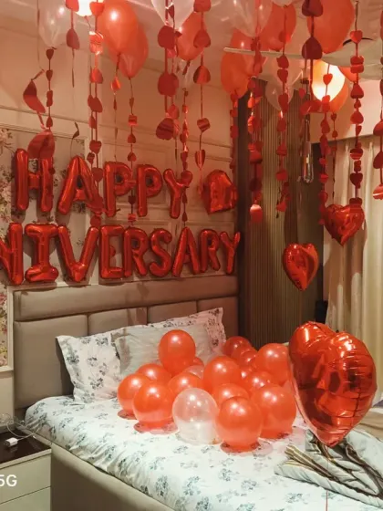 A full view of a romantic anniversary bedroom decoration. The room is filled with red heart-shaped balloons, hanging streamers, and large "Happy Anniversary" foil balloons above the bed.