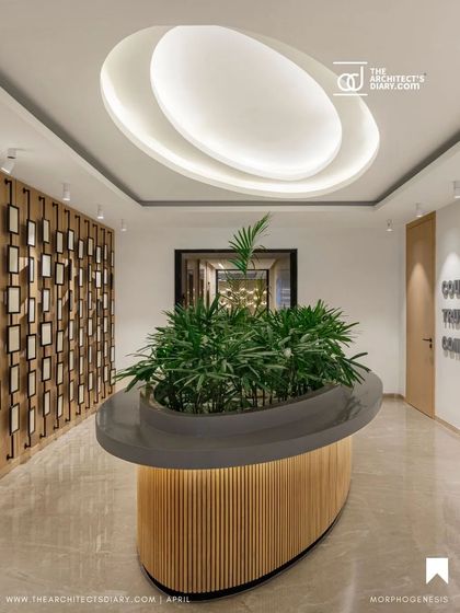 The reception area at Adani Realty features a central planter with lush greenery, with a layered ceiling light that emulates the planter's form. This biophilic element serves as a natural focal point and improves the indoor environment.