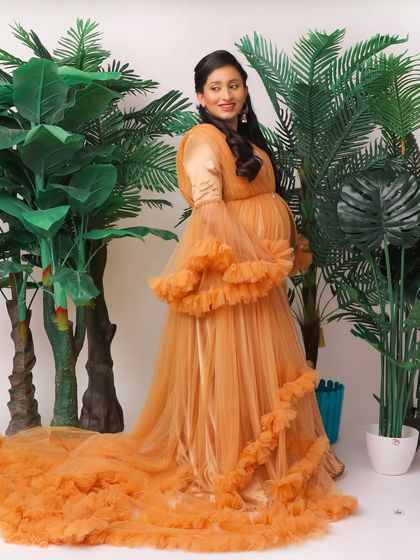 A beautiful full-length portrait of a mother-to-be in a ruffled orange gown. The tropical plant props in the studio add a fresh, vibrant feel to the shot.