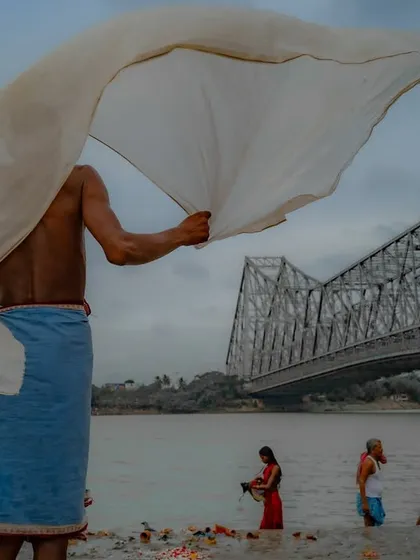 A man dries a large white cloth on the banks of the Hooghly river, with the Howrah Bridge in the background. The flowing cloth creates a sense of movement and freedom.