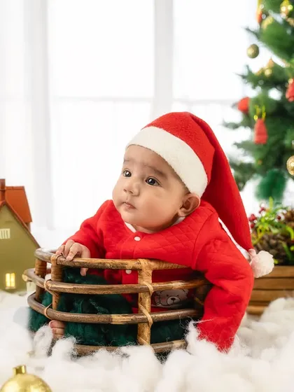 This baby Santa looks thoughtfully towards the Christmas tree, perhaps waiting for his reindeer. The miniature house adds a charming touch to the scene.