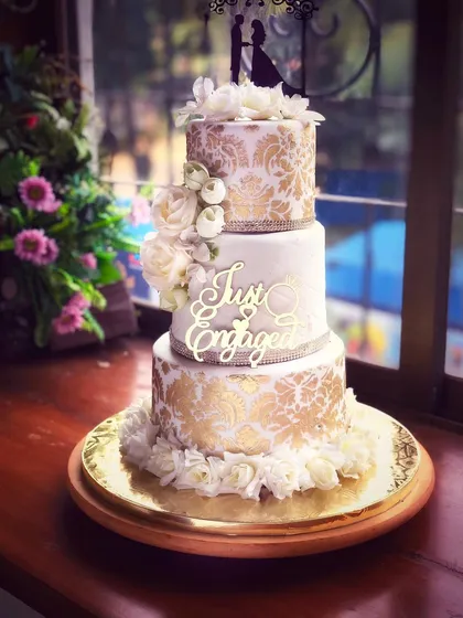 The stunning gold and white engagement cake displayed in natural light, showcasing its height and beautiful floral arrangement.