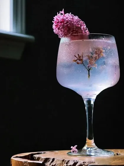A magical-looking gin and tonic with a color-changing infusion, garnished with a sprig of lilac. The presentation, with a tree-like flower inside, creates a miniature world within the glass.