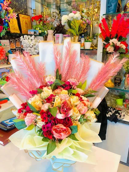 A large, luxurious bouquet of artificial flowers. This arrangement includes peonies and roses in shades of pink and cream, accented with dramatic pink pampas grass.