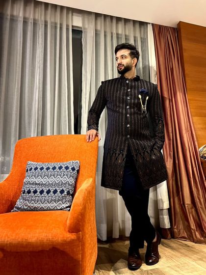 A full-length view of a dark, pinstriped Indo-Western sherwani. The outfit is accessorized with a stylish brooch, showcasing a modern approach to traditional groomswear.