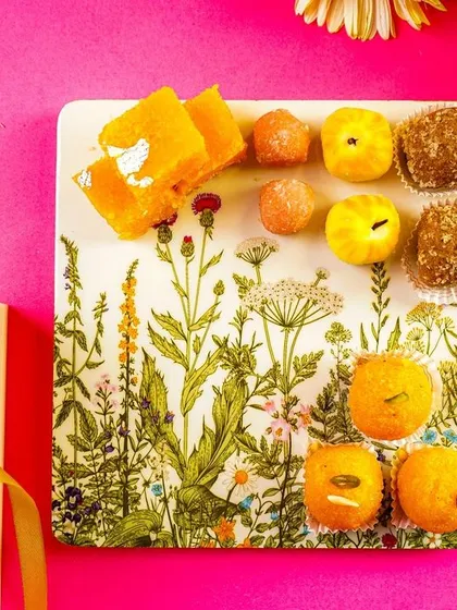 A festive product shot featuring a branded gift box and a platter of assorted Indian sweets.