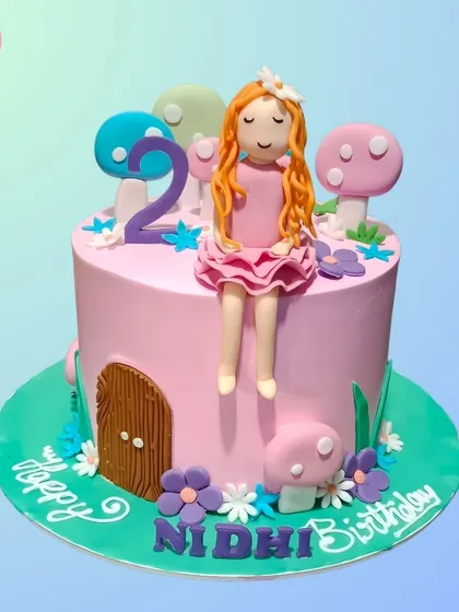A whimsical fairy-themed cake for a second birthday. It features a little fairy sitting on top of a mushroom house, surrounded by smaller mushrooms and flowers.