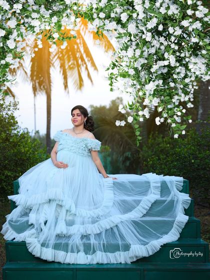 A dreamy portrait of a couple on green steps, surrounded by white blossoms. The mother-to-be's light blue ruffled gown adds to the ethereal feel.