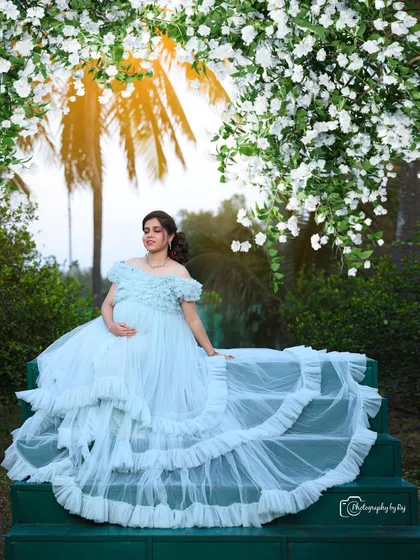 A dreamy portrait of a couple on green steps, surrounded by white blossoms. The mother-to-be's light blue ruffled gown adds to the ethereal feel.