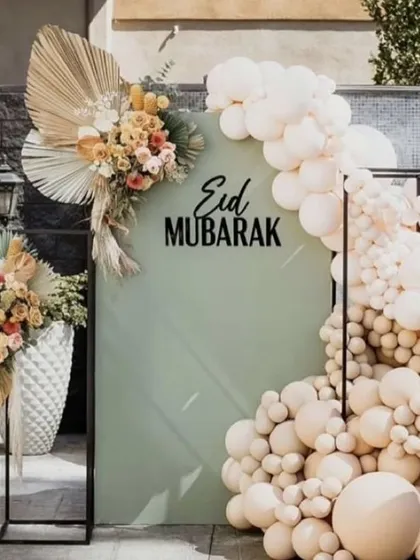 A modern and chic "Eid Mubarak" photo booth, with a sage green backdrop, a neutral-toned balloon arch, and dried floral arrangements.
