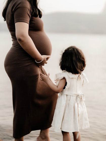A quiet moment between a mother and her first child. The little girl reaches out to touch her mom's belly, connecting with her new sibling by the peaceful water's edge.