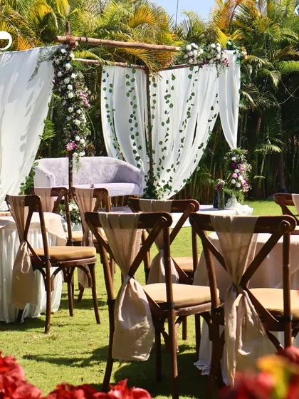An outdoor Roka ceremony setup featuring a rustic wooden mandap draped with sheer white fabric. The cross-back chairs with linen ties and surrounding greenery created a romantic, garden-party atmosphere.