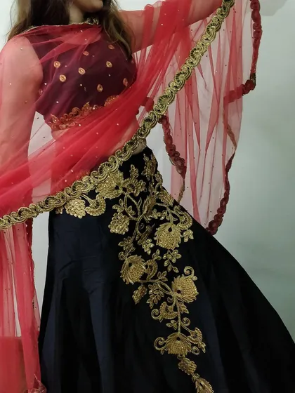 A detailed shot of the black lehenga, focusing on the heavy gold floral embroidery that climbs up the side of the skirt. The contrasting red dupatta adds a pop of color.