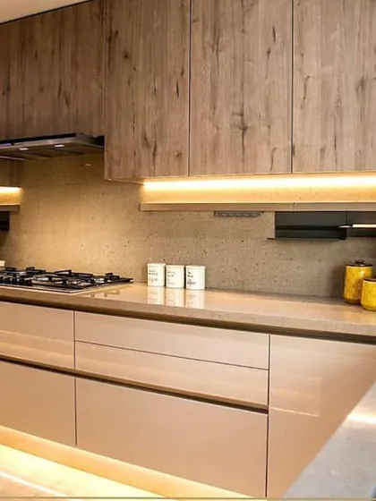 A warm and inviting kitchen featuring a mix of wood-grain and beige laminate finishes. The strip lighting under the cabinets and along the floor adds a modern, floating effect.
