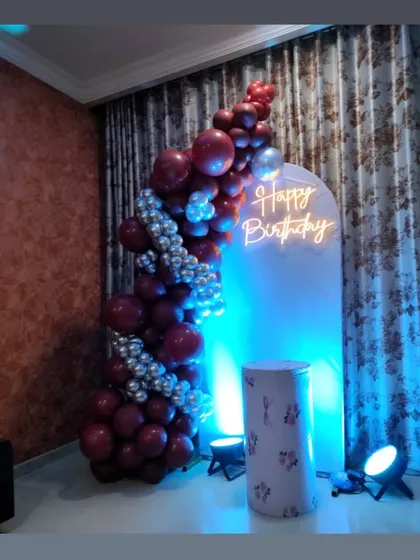 A stylish and modern backdrop featuring a maroon and silver balloon half-arch against a white panel with a neon sign. The blue uplighting adds a cool, contemporary feel.