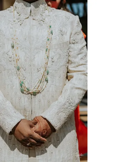 A detail shot of the groom's wedding attire. The focus here is on the elegant, multi-strand pearl and emerald necklace, which adds a touch of royal sophistication to his embroidered sherwani.