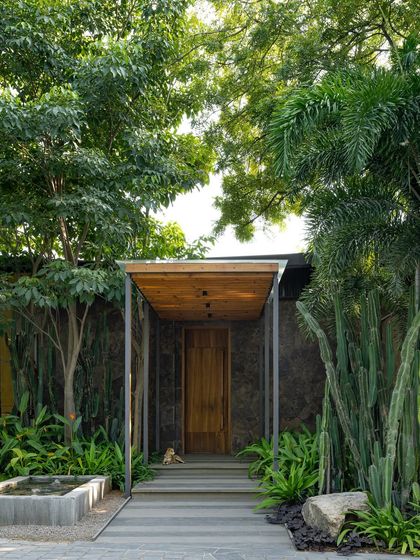 The entrance to the KEUS commercial building, featuring a modern wooden canopy and door. The pathway is flanked by tall cacti and other plants, creating a unique desert-modern aesthetic.