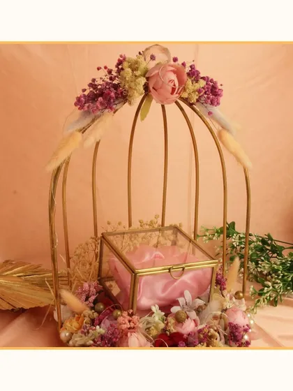 A beautiful engagement ring platter featuring a glass box nestled inside a decorative golden birdcage with floral accents. This design adds a touch of whimsy and elegance to the ceremony.
