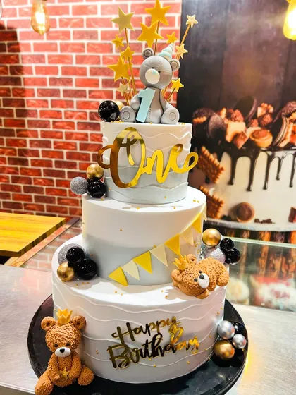 A grand three-tier teddy bear cake for a first birthday, decorated in a stylish black, white, and gold theme.