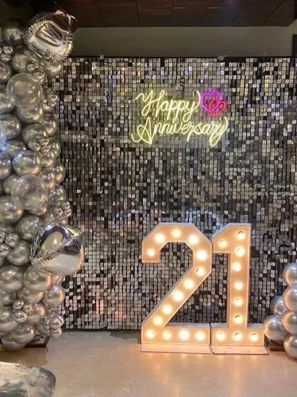 A chic 21st-anniversary party with a silver shimmer wall, a 'Happy Anniversary' neon sign, and '21' marquee numbers.