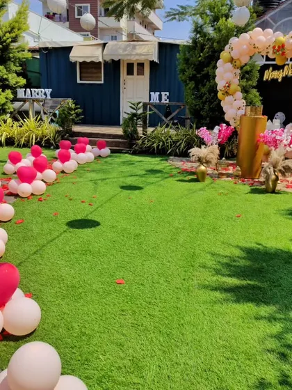 A playful and bright garden proposal setup. The pathway is lined with heart-shaped balloon clusters, leading to a large heart-shaped balloon arch in pastel pink and white, perfect for a cheerful and lovely surprise.