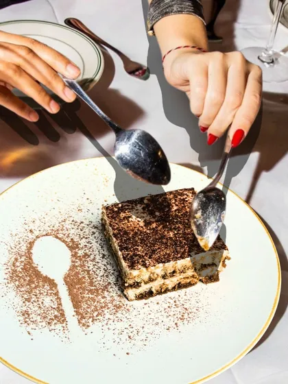 Pure indulgence. Two spoons digging into a slice of our classic Tiramisu, with a playful cocoa powder stencil on the plate.