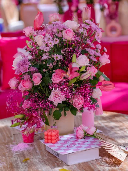 A vibrant floral centerpiece on a lounge table, with a Dior book and a bubble candle adding a touch of modern luxury. The hot pink velvet cushions in the background tie the whole look together.