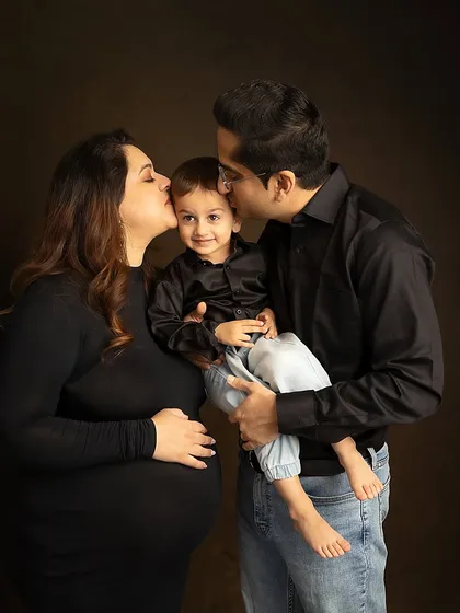 A classic family portrait against a dark background, filled with love and affection as both parents kiss their son.