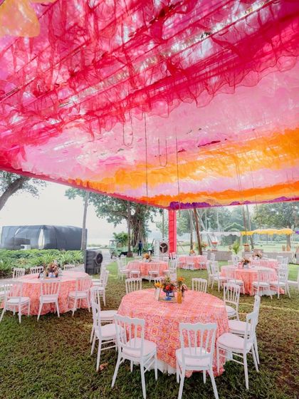A wide view of an outdoor event space with a ceiling of pink and orange drapes. The round tables are covered with patterned tablecloths, creating a cheerful and festive dining area.