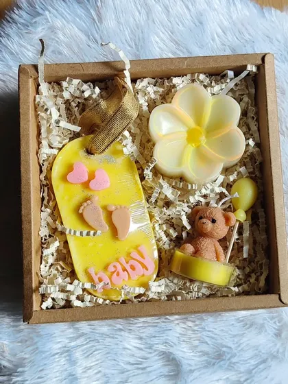 A close-up of the yellow baby shower gift box, featuring a baby-themed wax sachet, a teddy bear candle, and a flower soap.