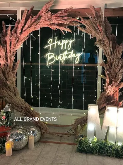 I design chic and trendy setups for any occasion. This rustic birthday decoration features a beautiful pampas grass wreath, a 'Happy Birthday' neon sign, marquee letter lights, and cozy, warm elements for a sophisticated celebration.