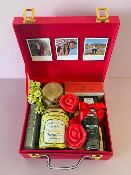 This red suitcase box is curated with a mix of self-care and gourmet items. It includes chamomile tea, a face mask, and chocolates, with polaroid photos for a personal touch.