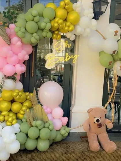 A lush and modern "Oh Baby" setup at an entrance. The organic balloon garland in shades of green, pink, and white is accented with a neon sign and a cute teddy bear prop.