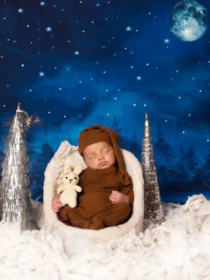 A sleeping newborn in a brown knit outfit, holding a tiny teddy bear. The baby is nestled in a soft white basket against our popular starry night backdrop.