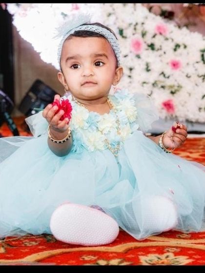 A sweet baby girl dressed in a beautiful blue party frock at her naming ceremony, sitting on a vibrant red carpet.