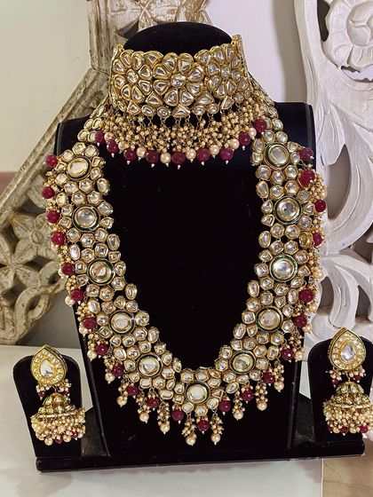 A full display of a classic bridal kundan set with ruby bead detailing. This includes a choker, a layered necklace, and matching jhumka earrings, available for rent or purchase.