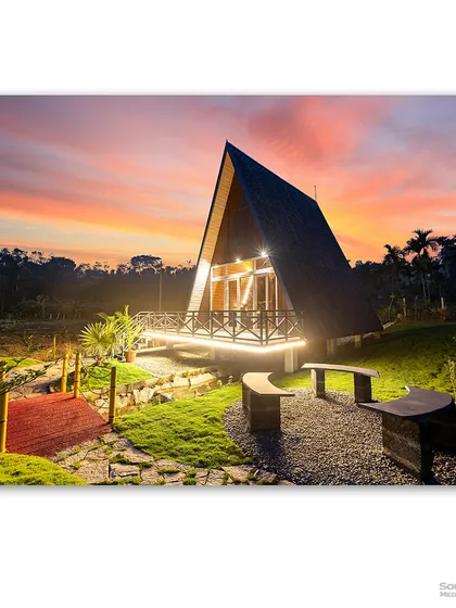 A cozy A-frame cabin glows warmly against a stunning sunset sky at Dewdrops, Sakleshpur. This image captures the magic of unique, sustainable architecture nestled in nature, highlighting the harmony between the built environment and its picturesque surroundings.