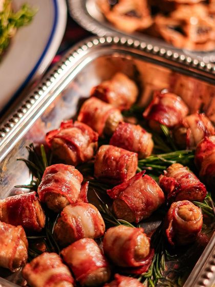 A tray of our pigs in a blanket, made with quality sausages and crispy bacon, resting on a bed of fresh rosemary. They are an irresistible part of our holiday catering menu.