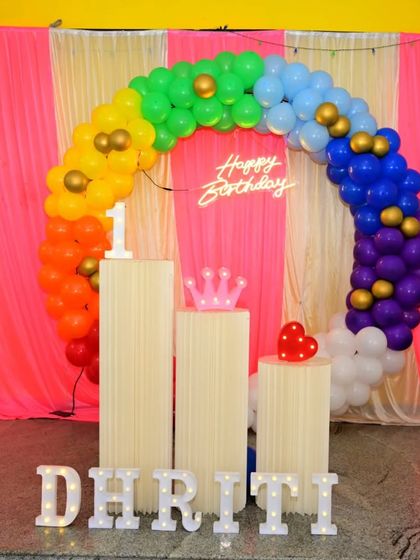 Another angle of the rainbow-themed first birthday party, showing the full circular balloon arch, neon sign, and marquee letters against a pink and yellow draped background.