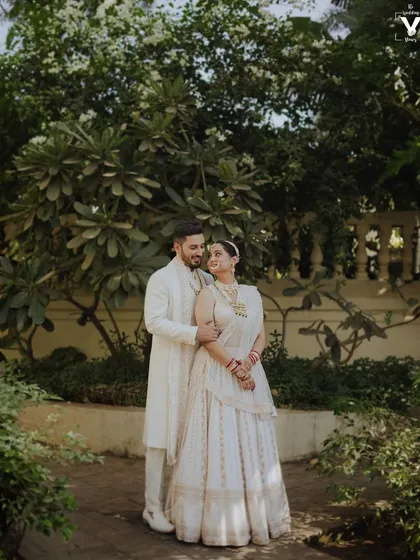 A classic portrait of the couple in matching white outfits, surrounded by lush greenery. Their elegant pose and the serene setting create a timeless and beautiful wedding photograph.