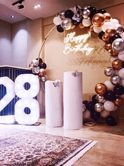 A stylish setup for a 28th birthday, featuring a black, white, and bronze balloon arch and marquee numbers.