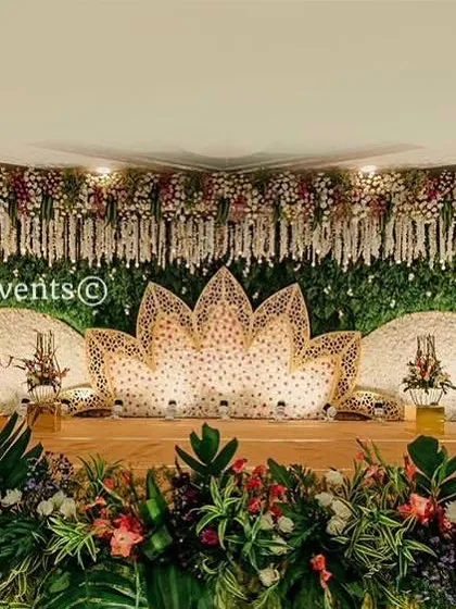 A grand reception stage featuring a stunning lotus-shaped seating area made of flowers. The backdrop is a mix of a green wall and hanging white florals, creating a wide, impressive visual.