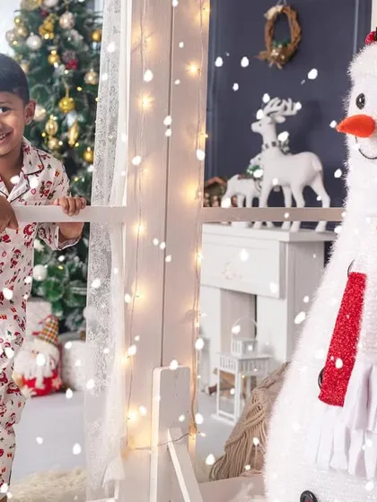 A young boy looks out the window at the falling snow, his face full of wonder, with a friendly snowman by his side.
