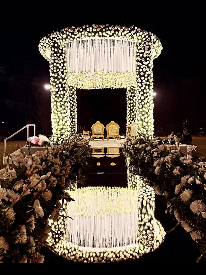 A dramatic night-time wedding stage decorated entirely with white flowers and lights, featuring a reflective aisle that creates a stunning mirror effect.
