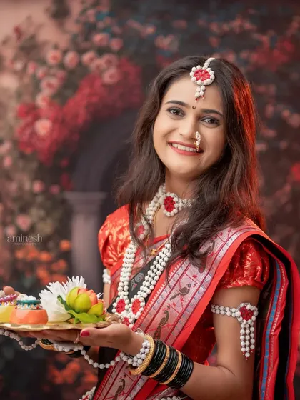 A smiling woman in a red and black saree holds a platter of tilgul against a floral backdrop. The soft lighting creates a warm and inviting festive photo.