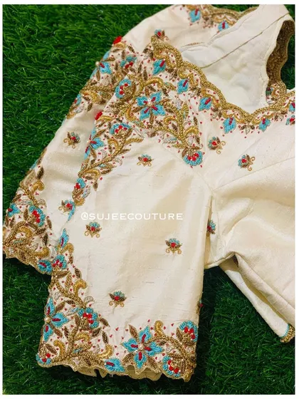 A beautiful cream-colored raw silk blouse. The design features intricate floral embroidery in turquoise, red, and gold zardozi, perfect for pairing with a bright-colored saree.